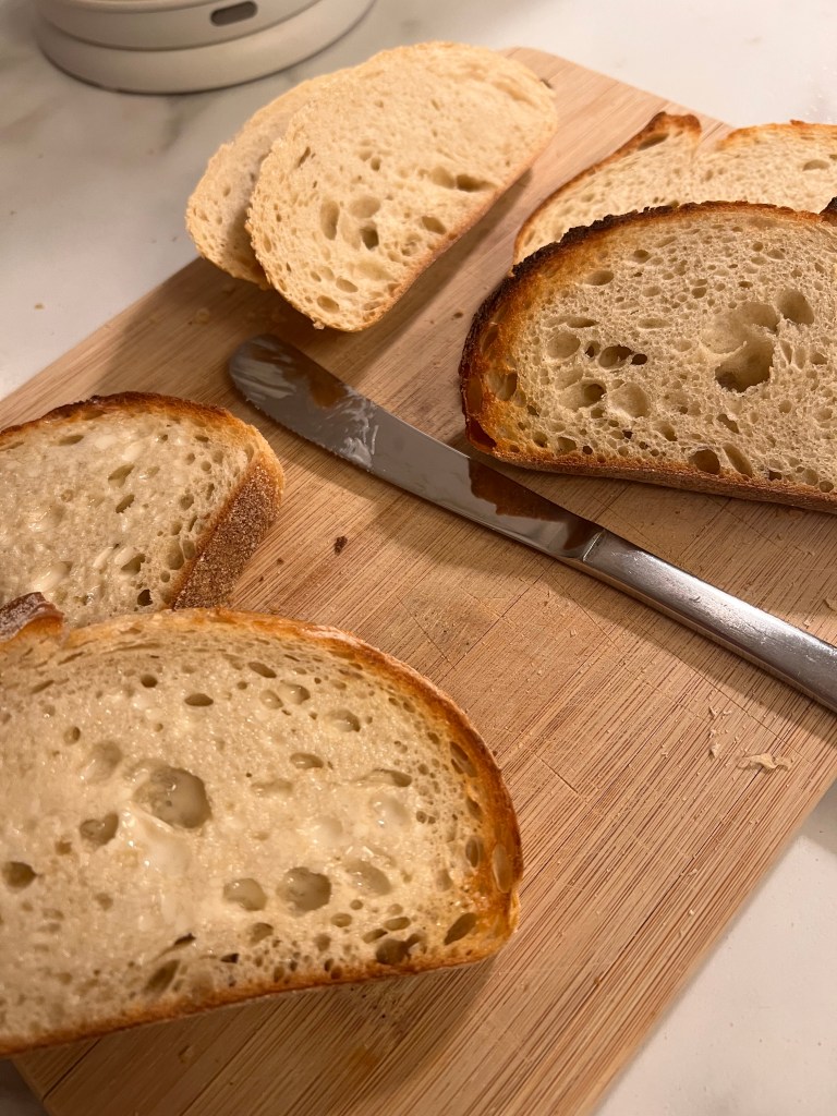 Costco’s Rustic Italian Bread loaf on a wooden board, sliced to show its thick crust and dense crumb.”